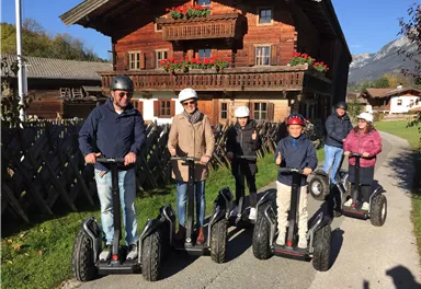 Eine Gruppe von sechs Personen fährt auf Segways in ländlicher Umgebung. Im Hintergrund ist ein traditionelles Holzhaupthaus mit Blumen zu sehen.