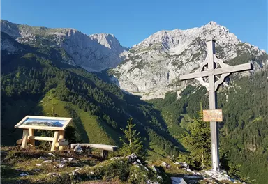 Eine schöne Berglandschaft mit hohen Gipfeln und grünem Wald. Im Vordergrund steht ein Holzkreuz und eine Informationstafel.