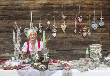 A woman in traditional clothing stands at a table full of handcrafted decorations. In the background, various jewelry pieces and hearts are hanging on a wooden wall.