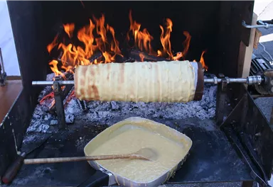 A wood stove with live flames, on which a spiral dough is being cooked. Next to it is a bowl of dough mixture.