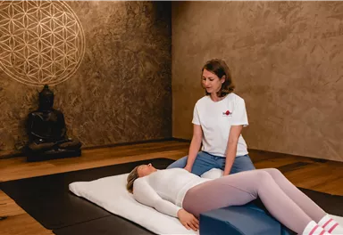 Eine Person führt eine Entspannungsbehandlung durch, während eine andere Person auf einer Liege liegt. Der Raum ist beruhigend gestaltet, mit einem Buddha und einer geometrischen Wanddekoration.