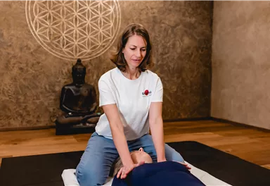 Eine Frau gibt eine Massage in einem ruhigen Raum. Im Hintergrund steht eine Buddha-Statue und ein Mandala ist an der Wand zu sehen.