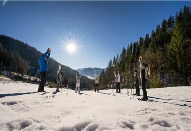 A group of people is doing a stretching exercise in the snow. The sun is shining brightly in the clear blue sky.