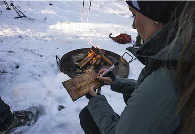 A person is sitting in the snow and writing on a piece of wood. In the background, a small fire is burning.