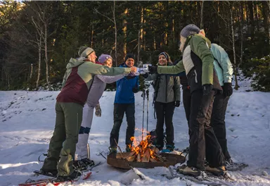 A group of six people stands in the snow around a campfire, toasting with drinks. In the background, trees and a wintry landscape are visible.