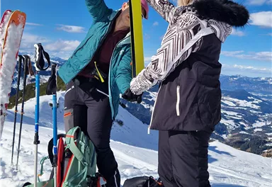 Two people are getting ready to ski in the snow. They are checking their equipment against a picturesque mountain landscape.