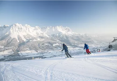 Zwei Skifahrer fahren die verschneite Piste hinunter. Im Hintergrund sind majestätische Berge und ein klarer Himmel zu sehen.