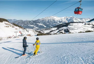 Two skiers are standing on a snowy slope and are looking at the impressive mountains. In the background, a gondola can be seen gliding over the landscape.