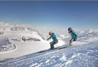 Zwei Skifahrer fahren einen verschneiten Hang hinunter. Im Hintergrund ist eine malerische Winterlandschaft zu sehen.