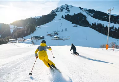 Two skiers are skiing down the slope, surrounded by snow-covered mountains. The sun is shining brightly in the blue sky.
