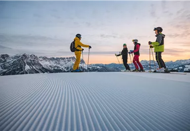 Eine Gruppe von Skifahrern steht auf einer frisch präparierten Piste in den Bergen. Die Aussicht zeigt schneebedeckte Gipfel und einen klaren Himmel bei Sonnenaufgang.