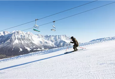 Ein Skifahrer fährt auf einer verschneiten Piste mit beeindruckenden Bergen im Hintergrund. Im Himmel sind Skilifte zu sehen.