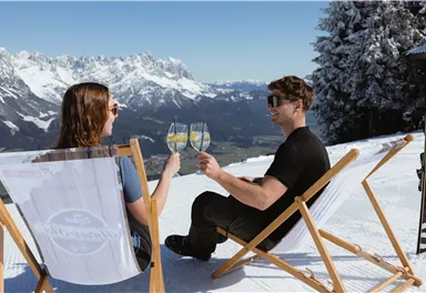 Ein Paar sitzt auf Liegestühlen im Schnee und stößt mit Gläsern an. Im Hintergrund sind schneebedeckte Berge und ein strahlend blauer Himmel zu sehen.