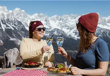 Zwei Frauen stoßen in den Bergen mit Gläsern an. Im Hintergrund sind schneebedeckte Berge und ein Tisch mit Essen zu sehen.