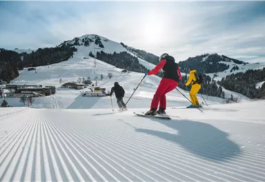Drei Skifahrer fahren die verschneite Piste hinunter. Im Hintergrund sind schneebedeckte Berge und eine klare Winterlandschaft zu sehen.