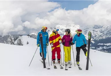 Eine Gruppe von vier Skifahrern steht auf einer verschneiten Piste. Im Hintergrund sind Berge und ein bewölkter Himmel zu sehen.
