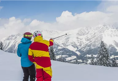 Zwei Skifahrer stehen im Schnee und blicken auf die beeindruckende Berglandschaft. Der Himmel ist teilweise bewölkt und die Umgebung ist von frisch gefallenem Schnee bedeckt.