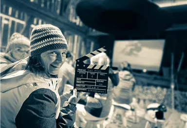 A cheerful person is holding a clapboard in a cinema or film room. In the background, there are other people and a large screen visible.