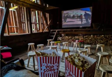 A cozy room with chairs and a screen in the background, showing a movie. In front, there are two bags of popcorn in striped cups.