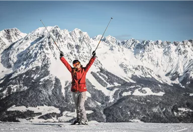 Eine glückliche Skifahrerin steht auf einem schneebedeckten Berg mit den Händen in die Luft. Im Hintergrund sieht man majestätische Berge und eine klare Winterlandschaft.
