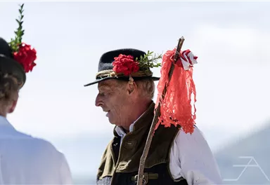 Ein Mann in traditioneller Kleidung mit einer blühenden Kopfbedeckung und einem Stock. Im Hintergrund sind unscharfe Silhouetten von anderen Personen zu sehen.