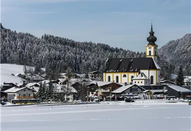 Eine schneebedeckte Landschaft mit einem charmanten Dorf und einer Kirche im Vordergrund. Im Hintergrund sind verschneite Berge und ein klarer Himmel zu sehen.