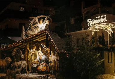 A festively lit Christmas nativity scene featuring figures of Mary, Joseph, and the baby Jesus. In the background, a decorated sign with the name of the place is visible.