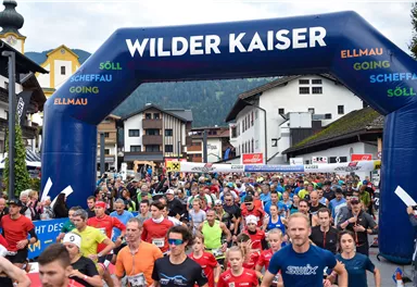 A large group of participants at the start of a race under a blue arch with the inscription "Wilder Kaiser". Many runners in colorful shirts are moving towards the start in a picturesque setting.