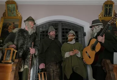 Eine Gruppe von vier Männern in historischen Kostümen steht vor einem Gittertor. Einer der Männer hält eine Gitarre und die anderen haben verschiedene Accessoires in der Hand.