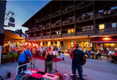 Eine belebte Straßenszene mit Musikern, die vor einem Holzgebäude auftreten. Viele Menschen sitzen draußen und genießen die Atmosphäre am Abend.