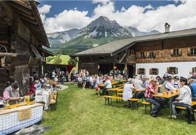A lively mountain festival with many people sitting at tables outdoors. In the background, picturesque mountains and traditional wooden houses can be seen.