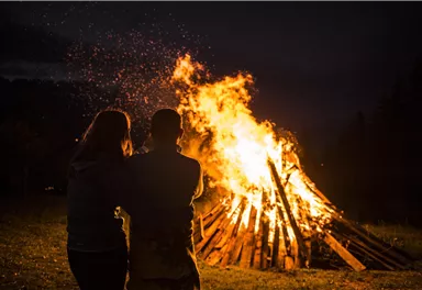 Ein romantisches Lagerfeuer bei Nacht. Zwei Personen stehen zusammen und betrachten die Flammen.