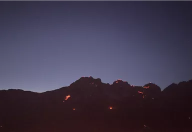 Ein gemütliches Berghaus aus Holz mit einer großen Terrasse. Umgeben von grünen Wiesen und Bäumen unter klarem Himmel.