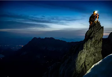 A climber stands on a high rock and inspects his gear. In the background, the mountainous landscapes and a clear sky at dusk are visible.