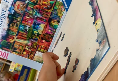 A child is puzzling with a colorful puzzle with many pieces. In the background, the packaging of the puzzle can be seen.