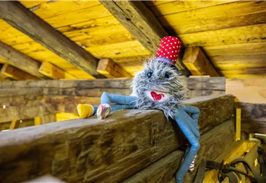 A funny plush toy with a red hat is sitting on a beam. It has a cheerful facial expression and colorful feet.