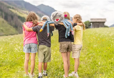 Four children are standing on a meadow and hugging each other. One of the children is holding a cuddly toy in the shape of a monster.