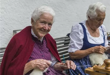 Two older women are sitting on a bench and doing handcrafting with wool. They are wearing traditional costumes and are smiling kindly.
