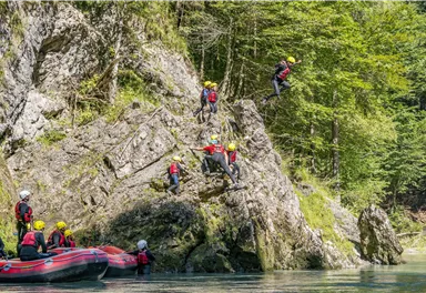 Eine Gruppe von Menschen springt von einem Felsen ins Wasser. Im Hintergrund sieht man grüne Bäume und ein Boot.