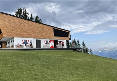 A modern building with a sloped roof structure stands on a meadow. In the background, there are mountains and a cloudy sky.