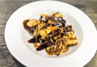 A delicious dessert on a white plate. It consists of fried bananas, garnished with chocolate sauce and powdered sugar.