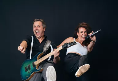 A musician with a guitar and a singer are happily posing in front of a neutral background. Both radiate energy and joy.