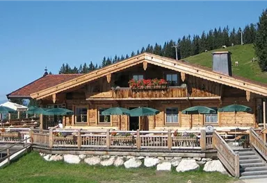 Ein gemütliches Berghaus aus Holz mit einer großen Terrasse. Umgeben von grünen Wiesen und Bäumen unter klarem Himmel.