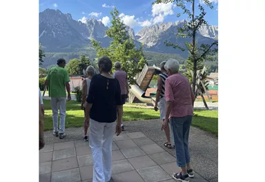 Eine Gruppe älterer Menschen spaziert in einer schönen Landschaft mit Bergen im Hintergrund. Der Himmel ist blau und die Umgebung ist grün.
