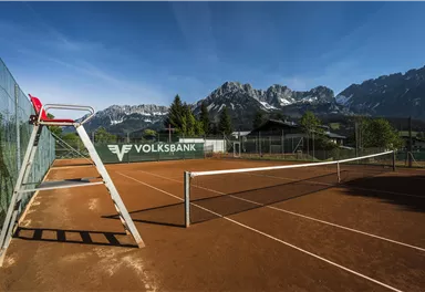 Ein Tennisplatz mit rotem Sand und einer imposanten Bergkulisse im Hintergrund. Die Sonne scheint und der Himmel ist klar.