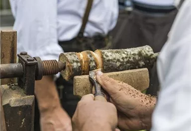 A craftsman is working on wood at a lathe. In the background, there are other people watching the process.