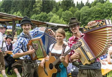 A cheerful group of musicians is playing at an outdoor festival. They are wearing traditional costumes and are surrounded by a picturesque forest landscape.