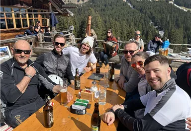 Eine Gruppe von sechs Männern sitzt an einem Tisch im Freien und genießt Getränke. Im Hintergrund sind Bäume und Berge zu sehen, die eine alpine Landschaft darstellen.