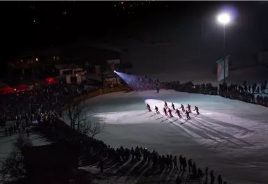 A winter event on a lit ice surface with several ice skaters. In the background, a large crowd of people and festive lights can be seen.