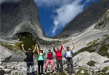Eine Gruppe von fünf Wanderern steht inmitten einer malerischen Berglandschaft. Sie feiern ihren Erfolg, indem sie die Hände in die Luft heben.